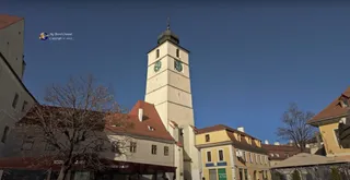 Un turn înalt cu ceasuri este situat între clădiri cu acoperișuri înclinate sub un cer senin.