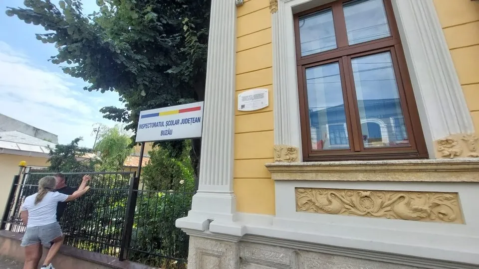 O femeie cu părul blond și tricou alb stă lângă un gard negru în fața unei clădiri galbene cu ferestre maronii.