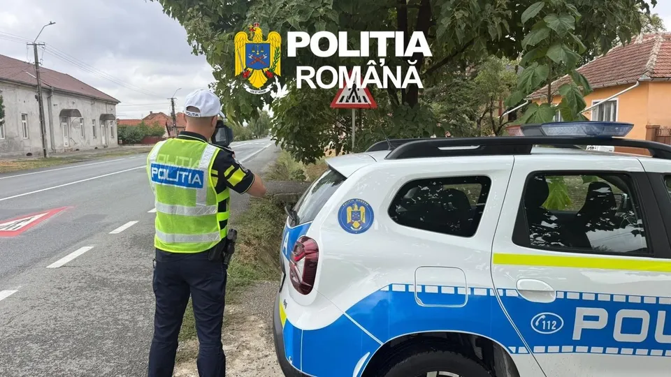 Un polițist cu vestă reflectorizantă stă lângă un autoturism alb cu marcaje albastre și galbenii pe marginea unui drum asfaltat.