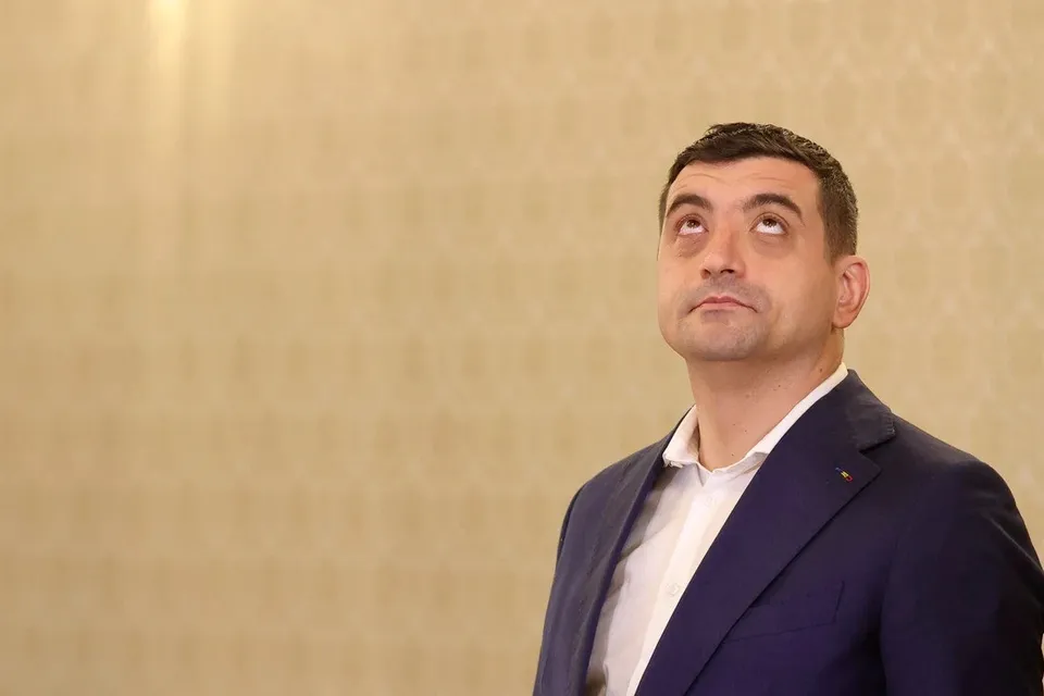 Un bărbat cu părul scurt și sacou albastru stă în picioare privind în sus în fața unui perete de culoare deschisă.