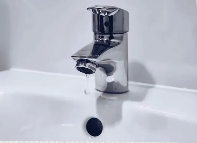 Un robinet metalic de culoare argintie este montat pe o chiuvetă albă, lăsând o picătură de apă să cadă din duză.