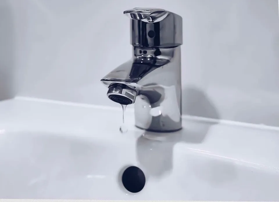 Un robinet metalic de culoare argintie este montat pe o chiuvetă albă, lăsând o picătură de apă să cadă din duză.