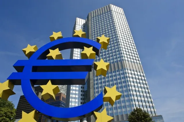Un simbol al monedei euro de culoare albastră cu stele galbene este plasat în prim-plan în fața unor clădiri înalte de birouri sub un cer senin.