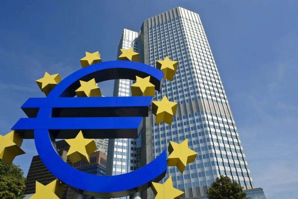 Un simbol al monedei euro de culoare albastră cu stele galbene este plasat în prim-plan în fața unor clădiri înalte de birouri sub un cer senin.