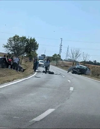 O mașină neagră este răsturnată pe marginea unui drum asfaltat, lângă care se află mai mulți oameni și un vehicul alb.
