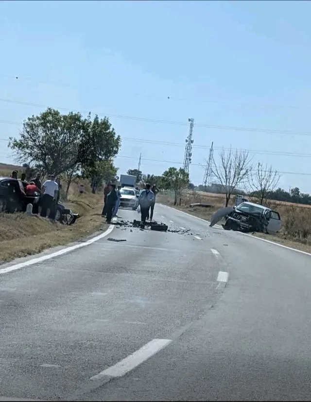 O mașină neagră este răsturnată pe marginea unui drum asfaltat, lângă care se află mai mulți oameni și un vehicul alb.