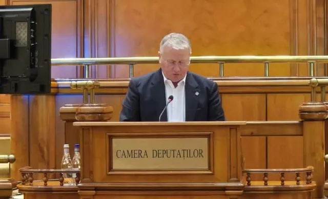 Un bărbat cu păr cărunt, îmbrăcat în costum, vorbește la un microfon, în spatele unui pupitru pe care scrie "CAMERA DEPUTAȚILOR".