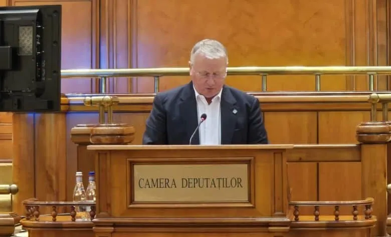 Un bărbat cu păr cărunt, îmbrăcat în costum, vorbește la un microfon, în spatele unui pupitru pe care scrie "CAMERA DEPUTAȚILOR".