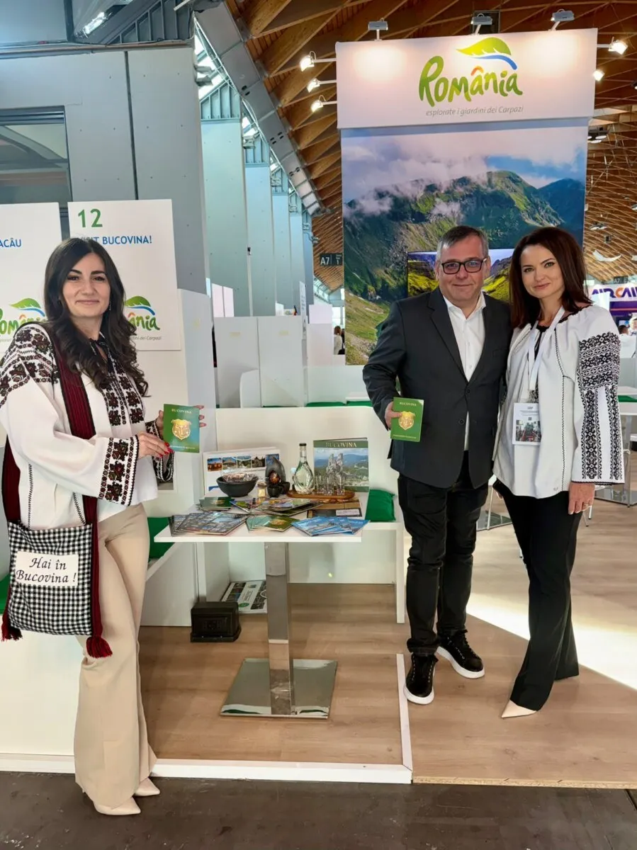 Trei persoane stau lângă un stand de expoziție cu un tabel alb, pliante și un panou cu textul România.