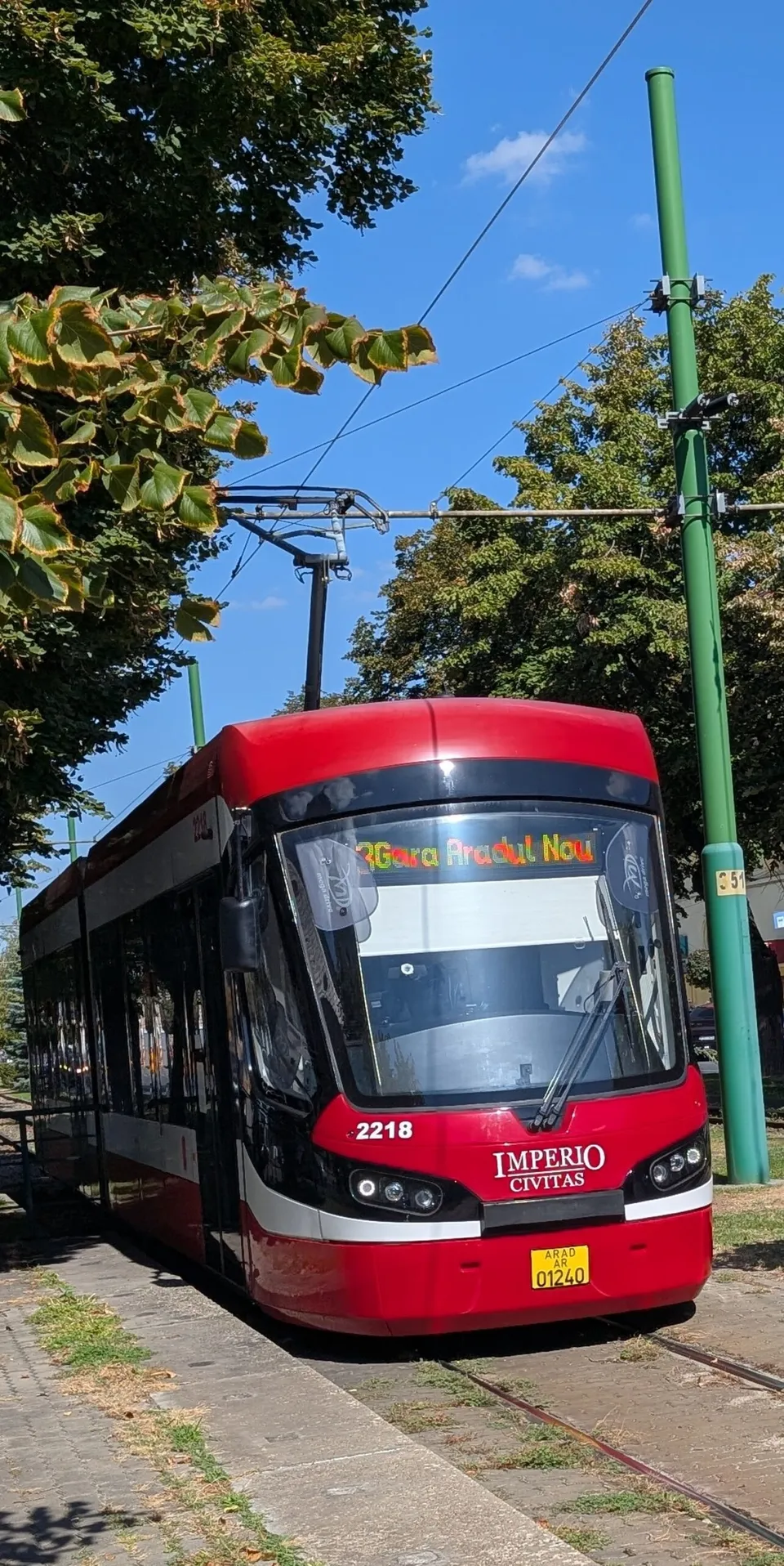 Un tramvai roșu și negru cu numărul 2218 circulă pe șine într-un mediu cu copaci și un stâlp verde sub un cer senin.