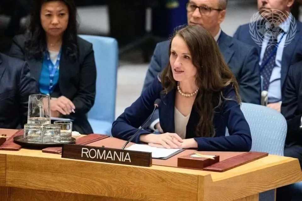 O femeie cu părul lung și sacou închis la culoare stă la o masă de conferință lângă un plac cu inscripția ROMANIA.