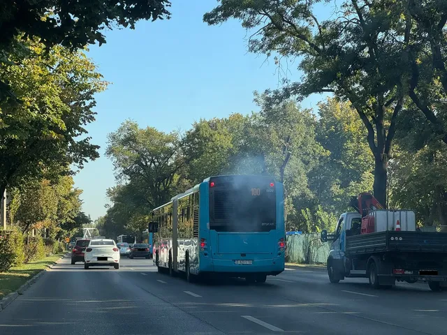 Un autobuz albastru de dimensiuni mari circulă pe un drum asfaltat, emanând fum de la partea din spate, în timp ce un camion este în apropiere.