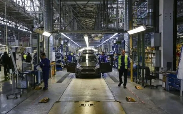 O mașină gri este poziționată pe o linie de asamblare într-o fabrică, înconjurată de muncitori și echipamente industriale sub lumini artificiale.