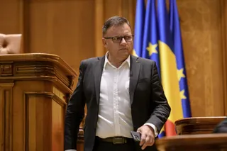 Un bărbat cu ochelari, purtând sacou negru și cămașă albă, stă în fața unor steaguri albastre și galbene într-o încăpere cu mobilier din lemn.