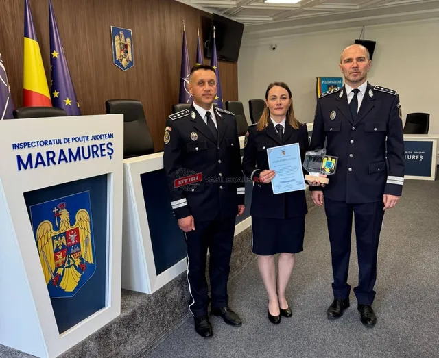 Trei persoane în uniforme oficiale stau în picioare într-o încăpere, în fața unui birou și al unui panou cu un blazon.
