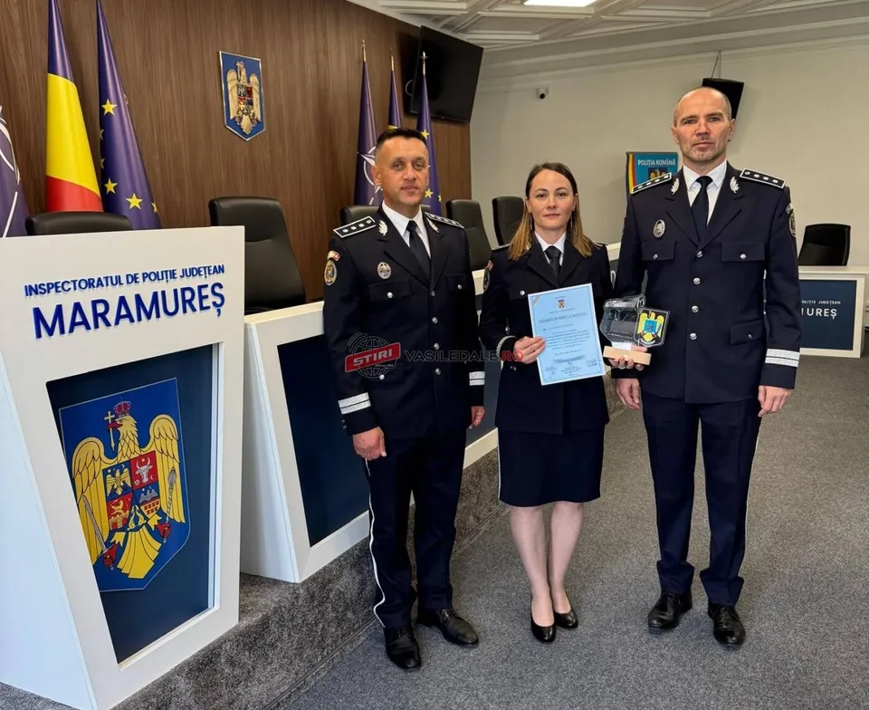Trei persoane în uniforme oficiale stau în picioare într-o încăpere, în fața unui birou și al unui panou cu un blazon.
