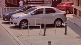 Două mașini parcate pe un trotuar de lângă un gard metalic alb și câțiva stâlpi metalici scurți.