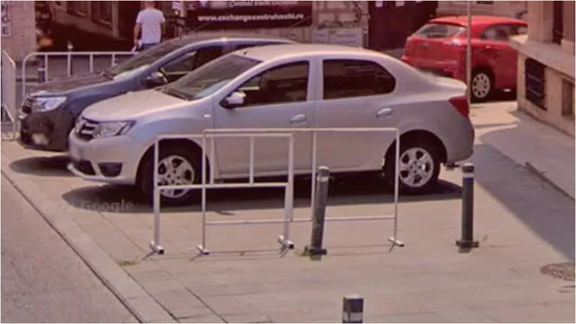 Două mașini parcate pe un trotuar de lângă un gard metalic alb și câțiva stâlpi metalici scurți.