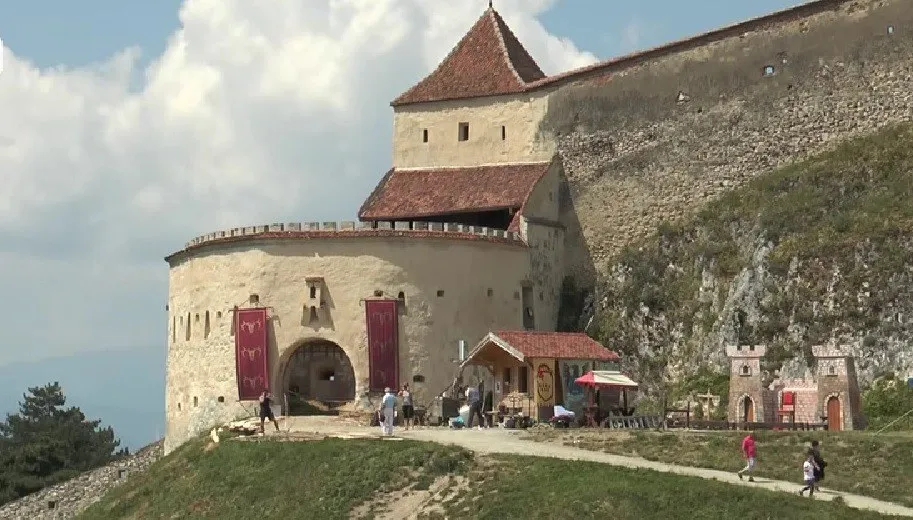 O cetate din piatră cu turnuri și ziduri înalte este situată pe un deal, cu câteva persoane mergând pe un drum de lângă clădire.