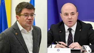 Un bărbat cu ochelari și sacou gri stă lângă un bărbat cu sacou negru, cămașă albă și cravată în timp ce acesta ține un pix.