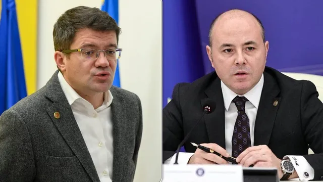Un bărbat cu ochelari și sacou gri stă lângă un bărbat cu sacou negru, cămașă albă și cravată în timp ce acesta ține un pix.
