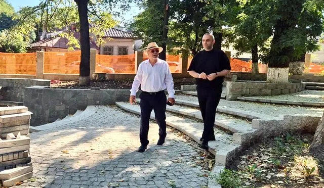 Un bărbat cu pălărie și cămașă albă merge pe un drum pavat lângă un alt bărbat îmbrăcat în negru.