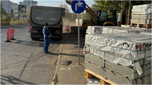 Un bărbat în costum de lucru albastru stă lângă un vehicul utilitar gri pe un drum asfaltat lângă stive de materiale de construcție.