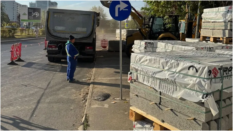Un bărbat în costum de lucru albastru stă lângă un vehicul utilitar gri pe un drum asfaltat lângă stive de materiale de construcție.