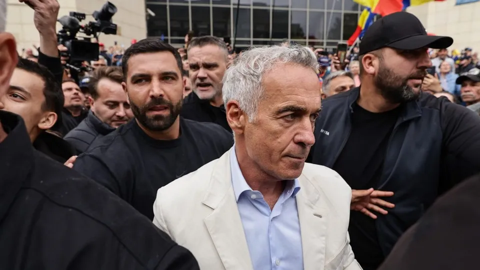 Un bărbat cu părul cărunt și sacou alb merge printre mai mulți bărbați în haine închise la culoare într-un spațiu exterior.
