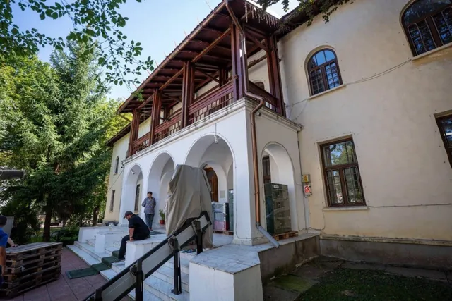 O clădire veche, cu un balcon din lemn, este văzută dintr-un unghi jos. Trei bărbați sunt pe trepte, iar în stânga jos se află paleți din lemn.