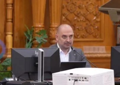 Un bărbat cu părul scurt și cămașă albă stă la o masă în fața unui monitor și a unui microfon într-o încăpere cu panouri de lemn sculptate.