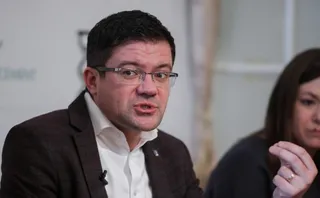 Un bărbat cu ochelari și sacou maro purtând o cămașă albă vorbește, în timp ce o femeie este parțial vizibilă în plan secund.