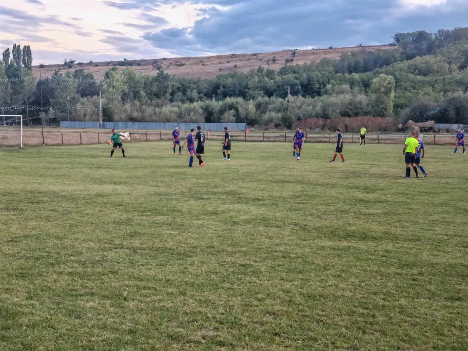Câțiva jucători de fotbal în echipamente colorate sunt împrăștiați pe un teren de gazon verde sub un cer înnorat.
