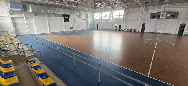 Sală sportivă interioară cu podea maro și albastră, linii albe, balustrade metalice și rânduri de scaune galbene și albastre.