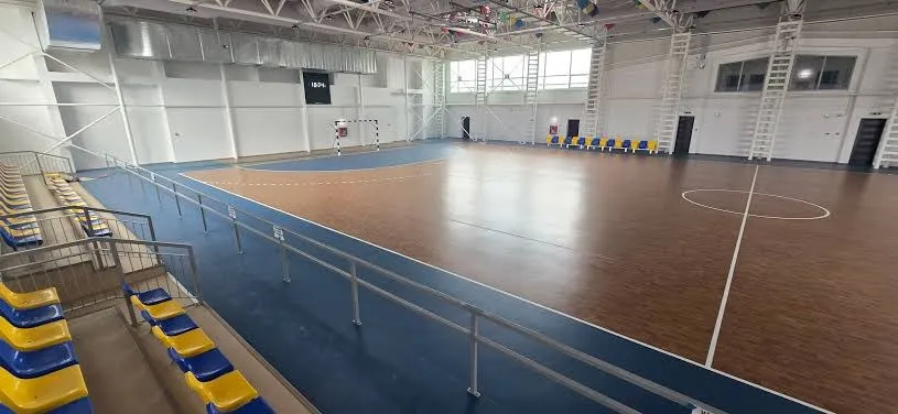 Sală sportivă interioară cu podea maro și albastră, linii albe, balustrade metalice și rânduri de scaune galbene și albastre.