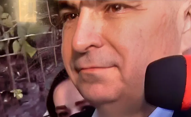 Un bărbat cu părul scurt este surprins în prim-plan, cu un microfon negru poziționat în fața feței sale.