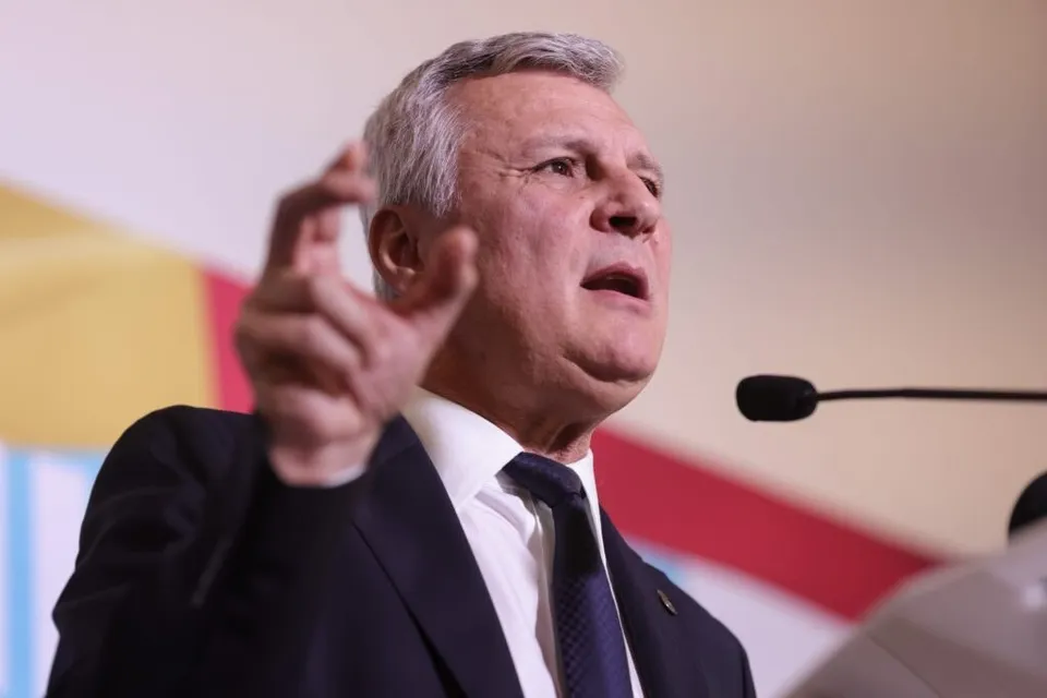 Un bărbat cu părul cărunt, purtând sacou închis la culoare, cămașă albă și cravată, ridică mâna stângă în timp ce se află în fața unui microfon.