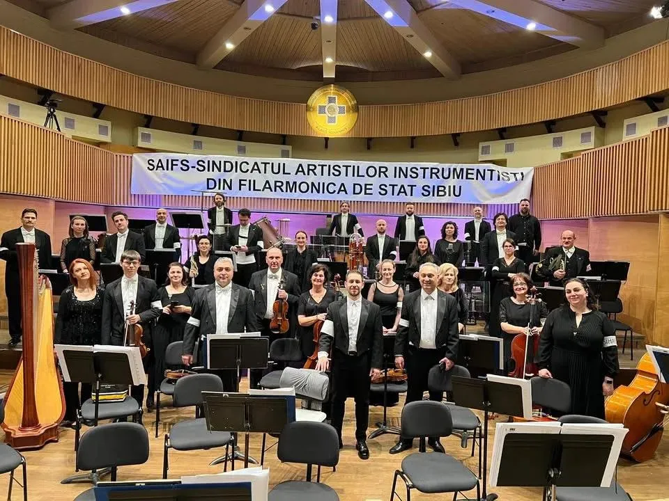 Un grup de muzicieni în haine negre stă pe o scenă de concert lângă harpă, violoncel și diverse instrumente muzicale.