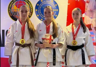 Trei fete purtând echipamente albe de karate și medalii cu pangalii roșii stau în picioare în fața unui panou colorat.