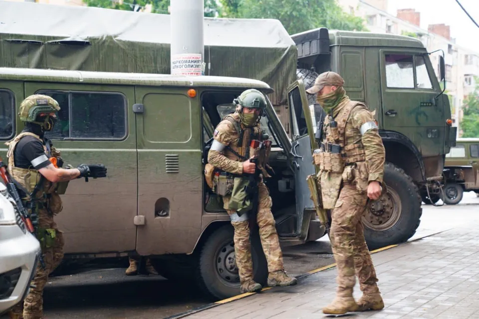 Trei soldați în echipament de luptă camuflat stau lângă un vehicul militar verde pe un drum urban.