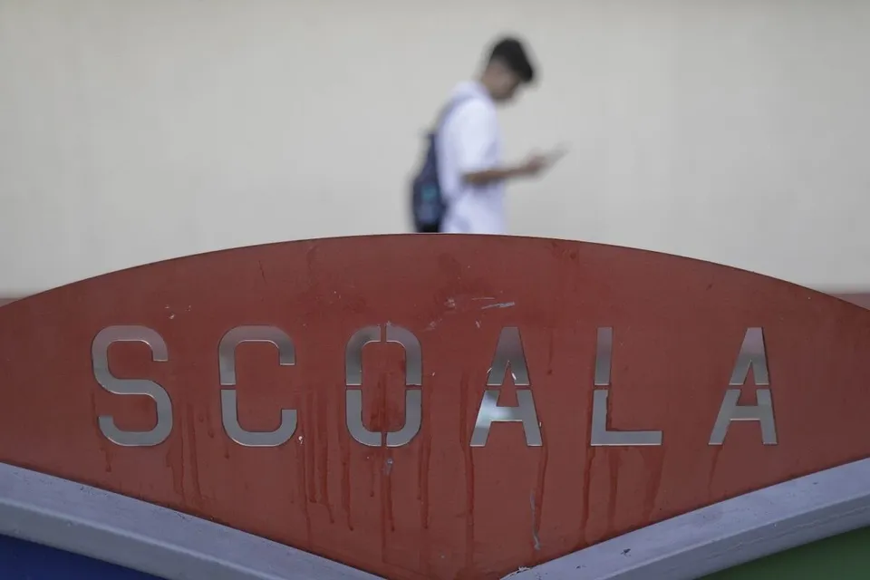 Un semn roșu cu textul SCOALA este în prim-plan, în timp ce un elev cu rucsac stă în fundal pe un fundal deschis.