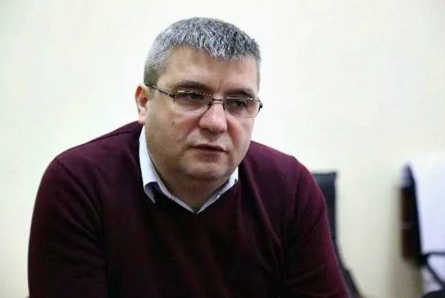 Un bărbat cu părul scurt și ochelari poartă un pulover bordeaux peste o cămașă albă.
