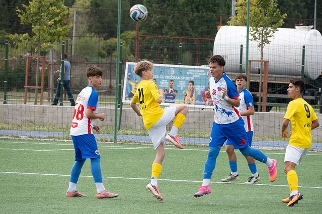 Cinci tineri jucători de handbal participă la un meci pe un teren de sintetice, având o minge în aer.