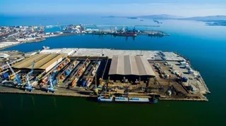 O vedere aeriană a unui port cu nave ancorate, macarale și clădiri industriale de depozitare situate pe malul unei ape calme sub un cer senin.