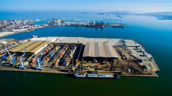O vedere aeriană a unui port cu nave ancorate, macarale și clădiri industriale de depozitare situate pe malul unei ape calme sub un cer senin.