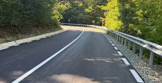 O porțiune de drum asfaltat cu linii albe marcate se întinde spre o curbă înconjurată de vegetație și de un parapet metalic.
