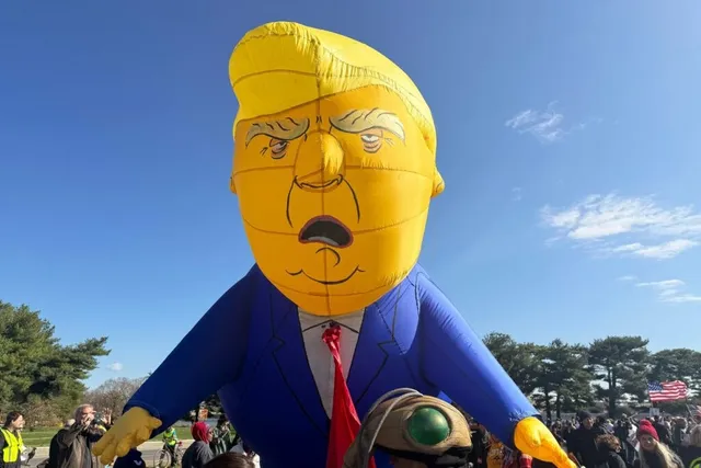 Un balon uriaș în formă de cap și trunchi de om cu păr galben, purtând costum albastru și cravată roșie, se ridică deasupra unei mulțimi sub cerul senin.