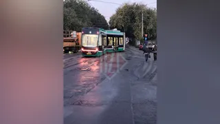 Un tramvai verde și alb stă pe șine într-o zonă urbană lângă un camion și câțiva oameni pe trotuar.