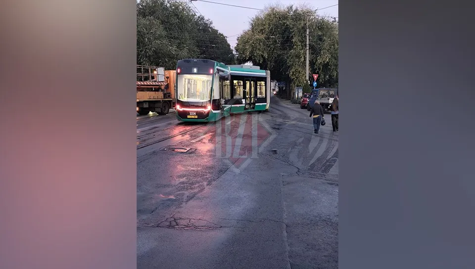 Un tramvai verde și alb stă pe șine într-o zonă urbană lângă un camion și câțiva oameni pe trotuar.