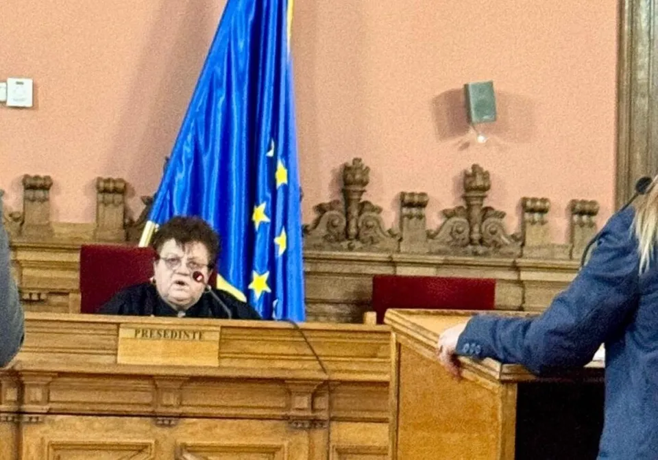 O femeie cu ochelari stă la un birou de lemn maro în fața unui steag albastru cu stele galbene.